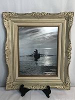 Framed black and white print depicting fisherman in boat on water under cloudy sky, shown on a black stand.