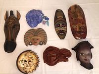 Top down view of 8 assorted masks including wood carved zebra, blue glass mask, Italian hand-painted ceramic sun and moon, and four wood ethnic style masks.