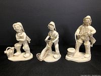 Three white ceramic fisherman figurines by Elfriede Meindl displayed side by side against black background, showing different poses and bases.