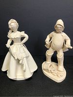 Two cream-colored ceramic figurines of a fisherman and his wife, standing side by side against a black background.