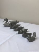 Six pewter duck figurines arranged in a line by size on white cloth background.