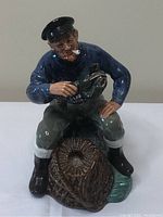 Front view of the Royal Doulton The Lobster Man figurine showing seated fisherman holding a lobster with a pipe in his mouth and basket beside him.