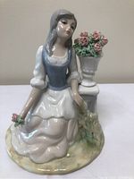 Front view of glossy porcelain figurine depicting a young woman in period attire kneeling beside a pedestal with floral vase.