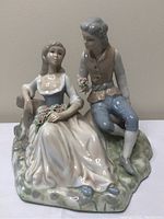 Front view of porcelain figurine of young couple seated on a base resembling grass and rock, showing both figures clearly with detailed facial features and clothing.