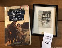 Softcover book titled The Old Curiosity Shop by Charles Dickens alongside a framed signed color sketch of a street scene showing 'The Old Curiosity Shop'.