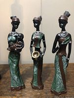 Front view of three standing figurines of African women, each in different poses and holding different items, dressed in green and brown with silver accents.