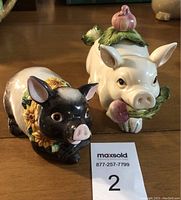 Photo showing white ceramic pig sugar dish with spoon and black ceramic pig potpourri container with sunflower decoration placed on surface with MaxSold lot number card.