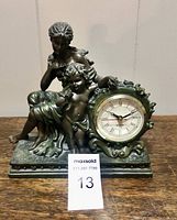 Front view of the mantel clock featuring mother and child figurine showing the full item on a wooden surface with identification tag number 13