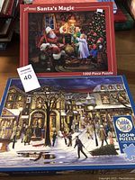 Photo showing the two boxed Christmas-themed puzzles side by side: 'Santa's Magic' 1000-piece puzzle on top and 'Tis the Season' 500-piece puzzle below.