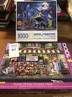 Two completed puzzles in their original boxes, displayed on a table with other puzzles in the background.