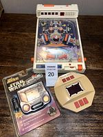 Photo showing three vintage electronic games on a wooden table: a white pinball game and two beige handheld LCD games, one still in original packaging.