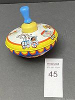 Front view of the metal spinning top showing circus-themed decorations with a blue handle and yellow collar.