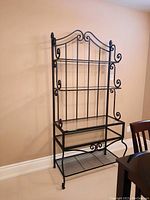 Full view of black metal bakers rack against wall showing decorative top and three glass shelves.
