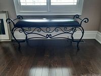 Front view of metal frame bench with black cushioned seat showing decorative scrollwork on legs and sides.