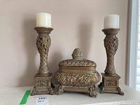 Two ornate carved candle holders flanking a decorative lidded box on a surface against a light wall.