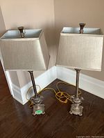 Pair of two matching gilded table lamps with beige fabric rectangular shades placed on wooden floor corner.