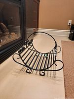Black metal log holder next to fireplace, showing curved base, horizontal slats, and side circular handles with scrollwork ends.