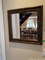 Square beveled glass mirror hanging on wall reflecting interior with staircase and dining area, showing overall condition and size.