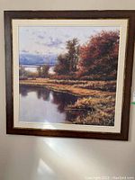 Framed print showing a landscape with a lake reflecting trees with fall foliage under a cloudy sky.