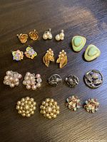 Nine pairs of vintage clip on earrings and a Scottish thistle brooch arranged on a dark surface, showing the variety of designs and styles.