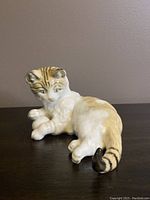 Porcelain cat figurine shown lying on its side on a dark surface against a neutral wall. The image clearly shows the coloring and details of the cat's fur, face, and curled paws.