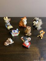 Photo showing collection of eight small porcelain animal figurines on a wooden surface against a grey wall, including mouse in blue outfit, fox, owl, red rose dog, skunk with fur, chipmunk, lying dog, and mouse with graduation cap.