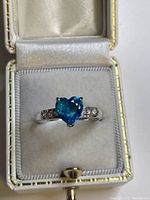Heart-shaped blue gemstone ring with clear gemstones on shoulders, displayed inside a white cushioned jewelry box with satin lining.