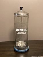 Full front view of the vintage Barbicide jar showing the metal lid, glass body with printed text, and rubber base.