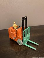 Front angled view of vintage orange and green metal forklift toy with seated driver figurine on black surface.