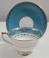 Side and top view of Aynsley tea cup with blue interior rim and gold filigree, matching saucer behind