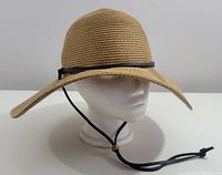Front and side angled view of a tan wide brim straw hat on a white mannequin head. The hat has a black leather band and an adjustable black chin strap with a wooden bead.