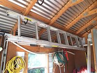 Aluminum extension ladder stored horizontally indoors on wooden supports under a corrugated roof, showing ladder length and rungs.