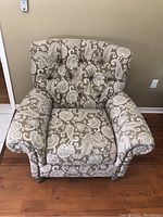Front angled view of cream and taupe paisley patterned armchair showing tufted back and rolled arms on wood floor.