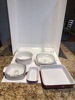 Seven baking dishes arranged on a white background on granite counter, two with glass lids. Various sizes, white ceramic and clear glass.