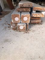 Stacked thick wooden beams painted grey, ends showing solid square cuts and natural wood grain with knots, placed on a concrete floor. Some dirt and debris visible on and around the beams.