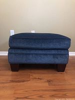 Front view of blue fabric ottoman showing cushioned top and wooden block legs on hardwood floor against beige wall.