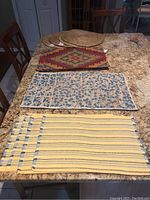 Four sets of placemats displayed on a granite countertop. Front set is yellow with blue and green stripes, second set has a teal floral pattern on beige/taupe background, third set is multicolor geometric pattern, fourth is woven circular natural fiber placemats.