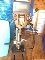 Full view of Ryobi drill press mounted on a metal stand showing base, column, table, motor, and feed handles.