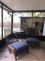 Photo showing two metal mesh chairs, two wicker ottomans with blue cushions, and a tapestry hanging on a hook in a screened patio enclosure.
