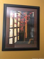 Close-up photo of framed wall art featuring a black vase filled with red flowers in front of a window. Shows details of frame and image.