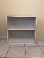 Front view of the white wooden bookcase showing two shelves, clean tiled floor background, beige wall.