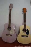 Front view of two acoustic guitars side by side against wall, pink on left and natural wood on right.
