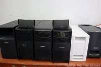 Photo showing six Bose media subwoofers in black and white colors and two smaller bookshelf speakers, front view showing Bose branding and grilles.