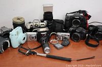 Photo showing all cameras, flash unit, and some camera accessories together on a white surface.