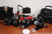 Overview of the camera lot showing Canon EOS 7D, Canon EOS Rebel, Nikon DSLR, a compact Canon camera, two camera bags, and camera straps.