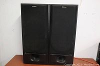 Front view of both speakers with fabric grills on, showing rectangular black wooden cabinets and front bass reflex port.