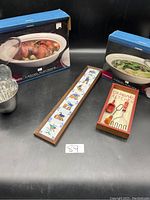 Photo showing full lot including two boxed Casual Elegance ceramic serving dishes, metal ice bucket, decorative tile wall art, and Grandma's Kitchen utensil decor set.