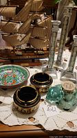 Photo showing wooden model ship with sails and rigging, porcelain bowl nearby, two mirrored candlesticks, two black and gold ashtrays, and green glass cat candle holder on lace tablecloth.