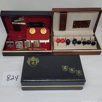 Overall view of two open cufflink and tie clip cases showing multiple pairs of cufflinks and several tie clips arranged inside, plus a closed black case with gold crest emblem.