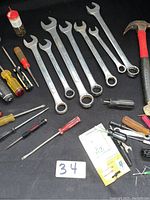 Photo showing 7 combination wrenches and a claw hammer with a red and black handle, assorted screwdrivers with different colored handles, small oil bottle on black cloth background.